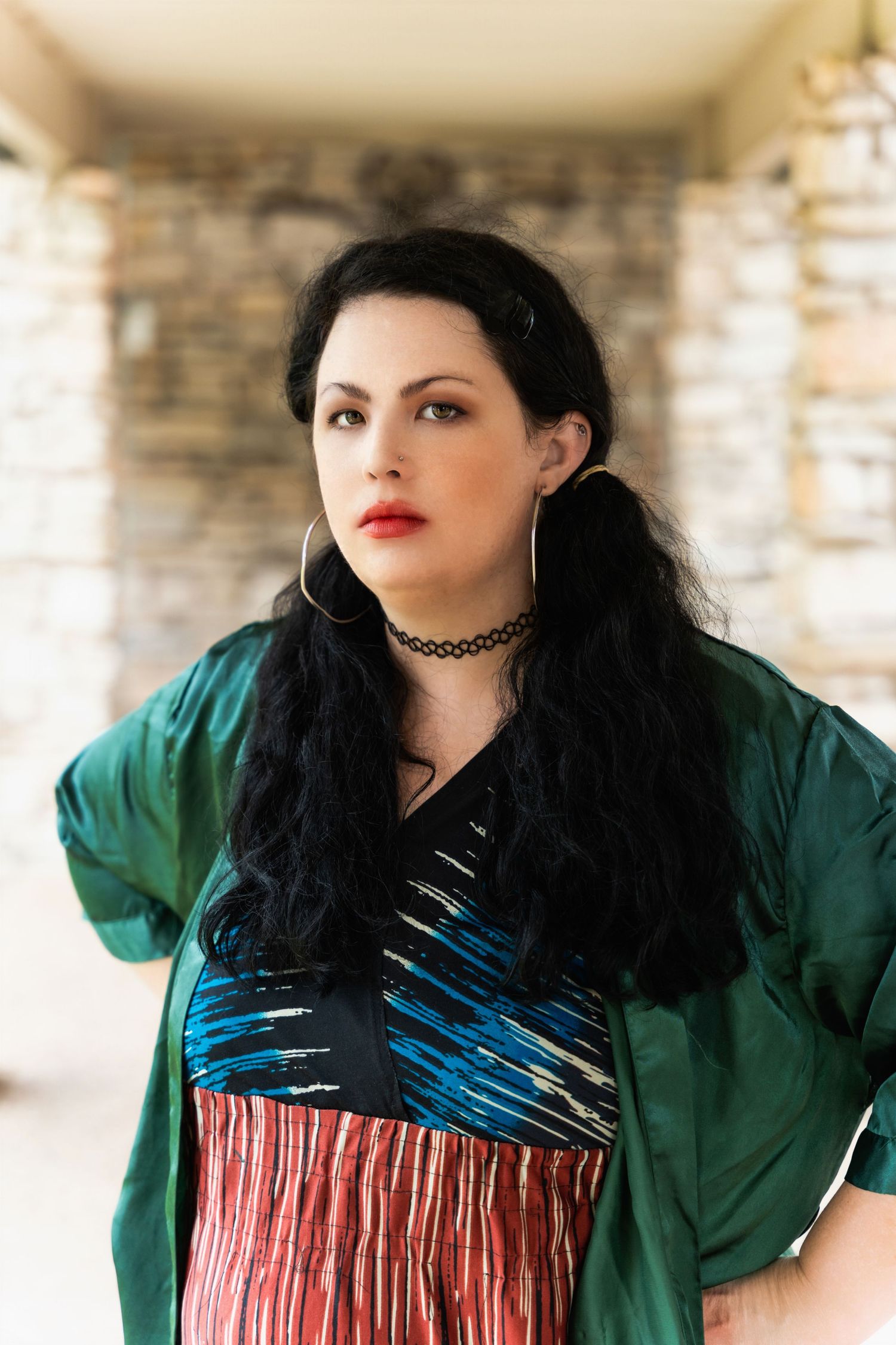 A portrait shot of someone wearing a green jacket and choker necklace with long dark hair against a brick wall background.