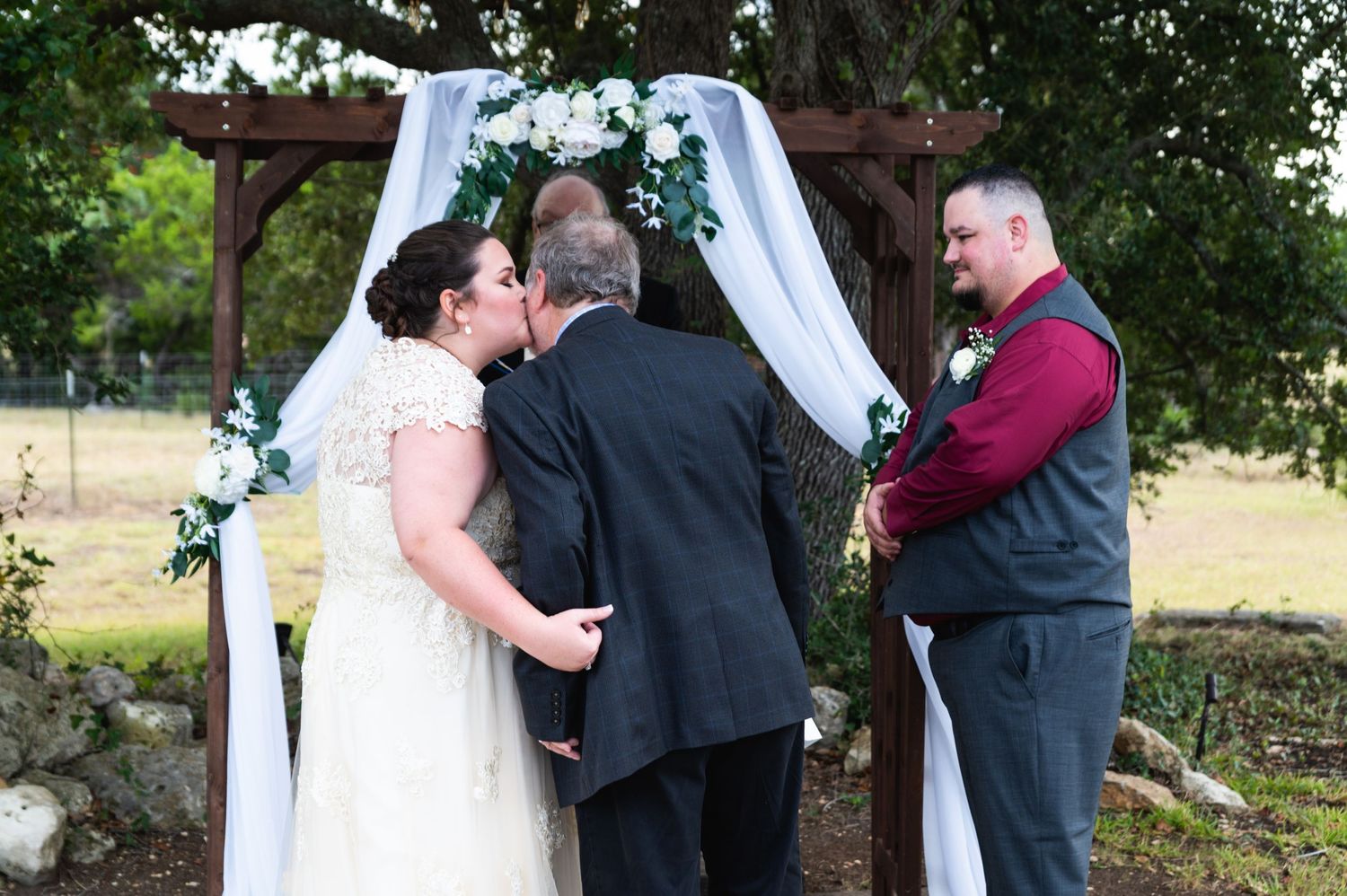 A wedding ceremony takes place under a wooden arch decorated with white fabric and greenery.