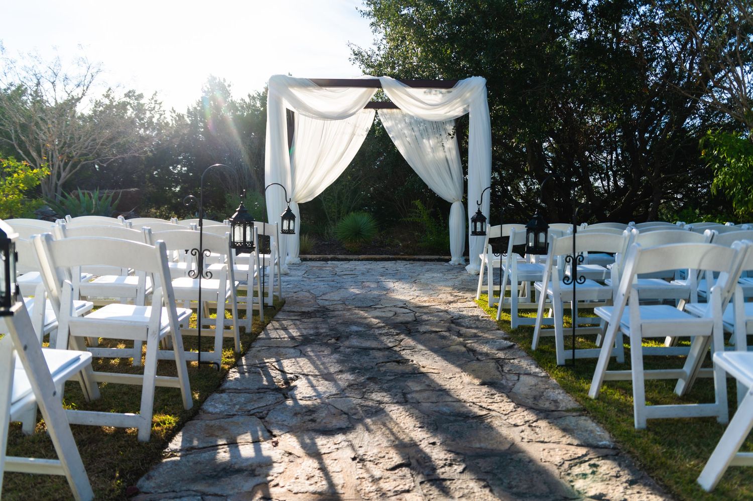 Outdoor wedding ceremony setup with white chairs and draped arch at sunset.