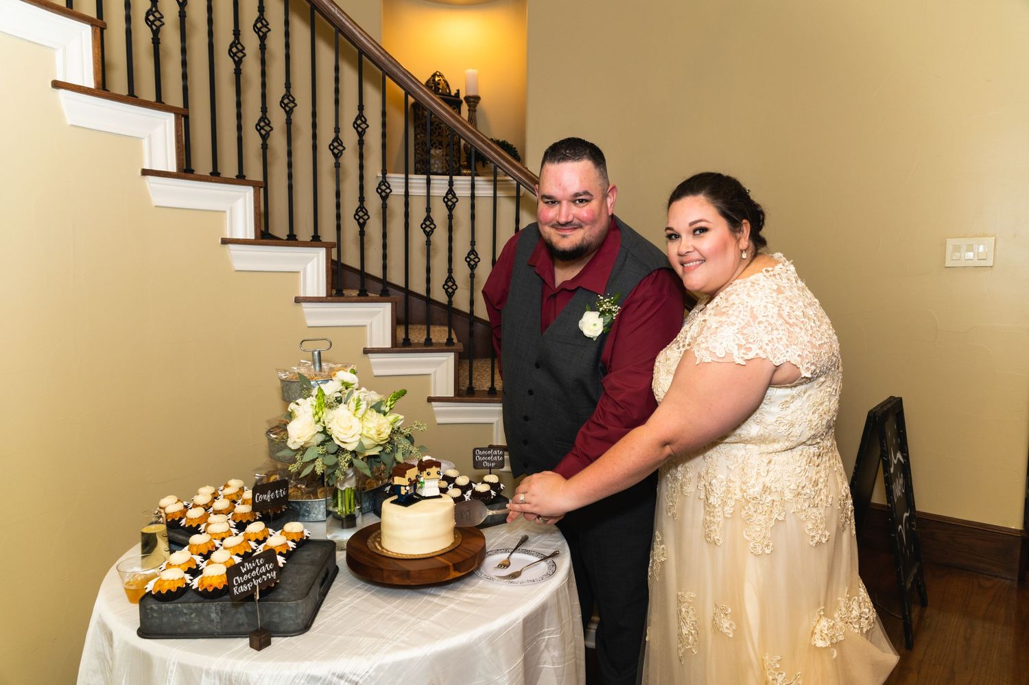 A couple cuts their wedding cake together at the reception, displayed on a table with cupcakes and floral decorations.