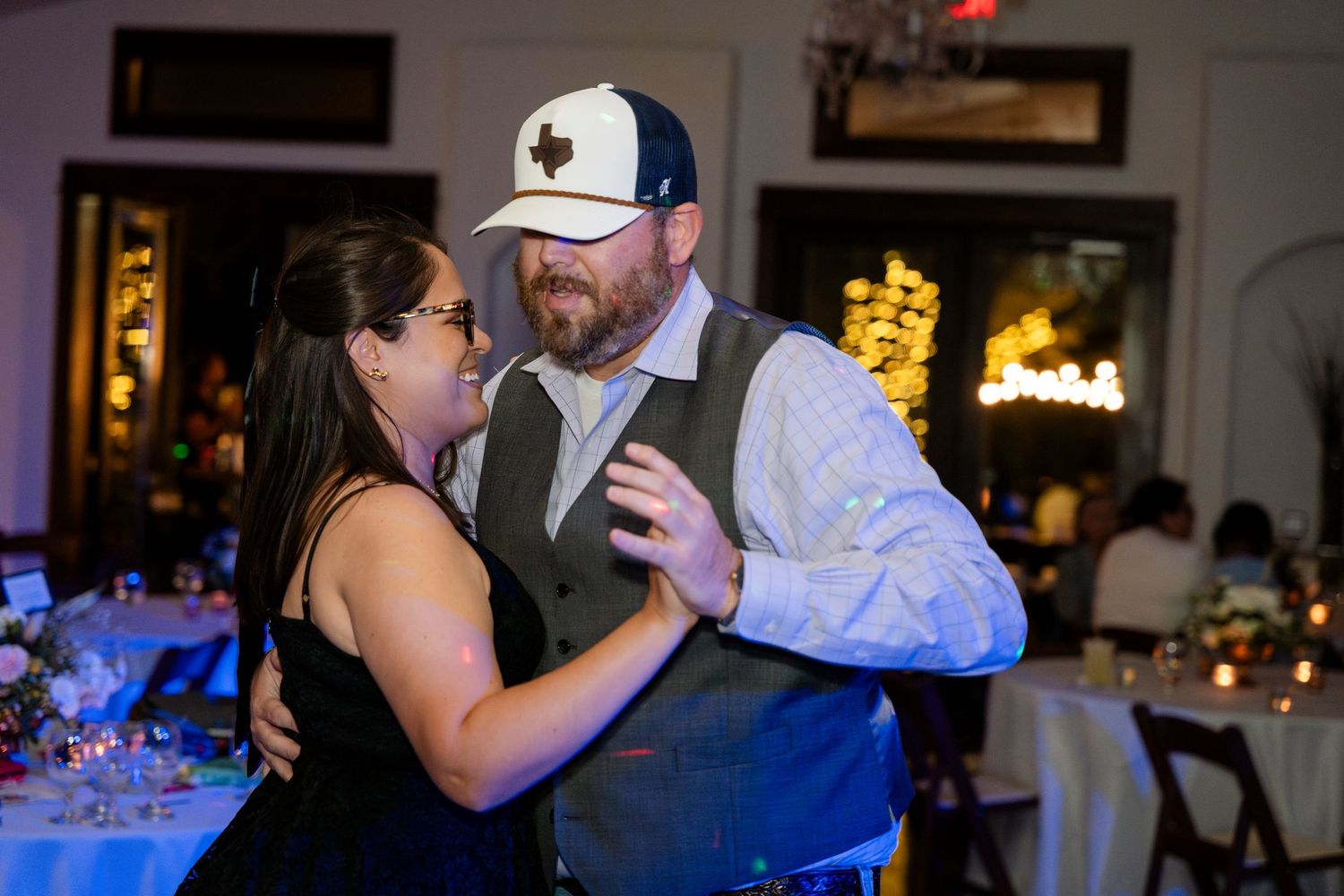 A couple shares a dance together at a wedding reception with blue lighting and table settings in the background.