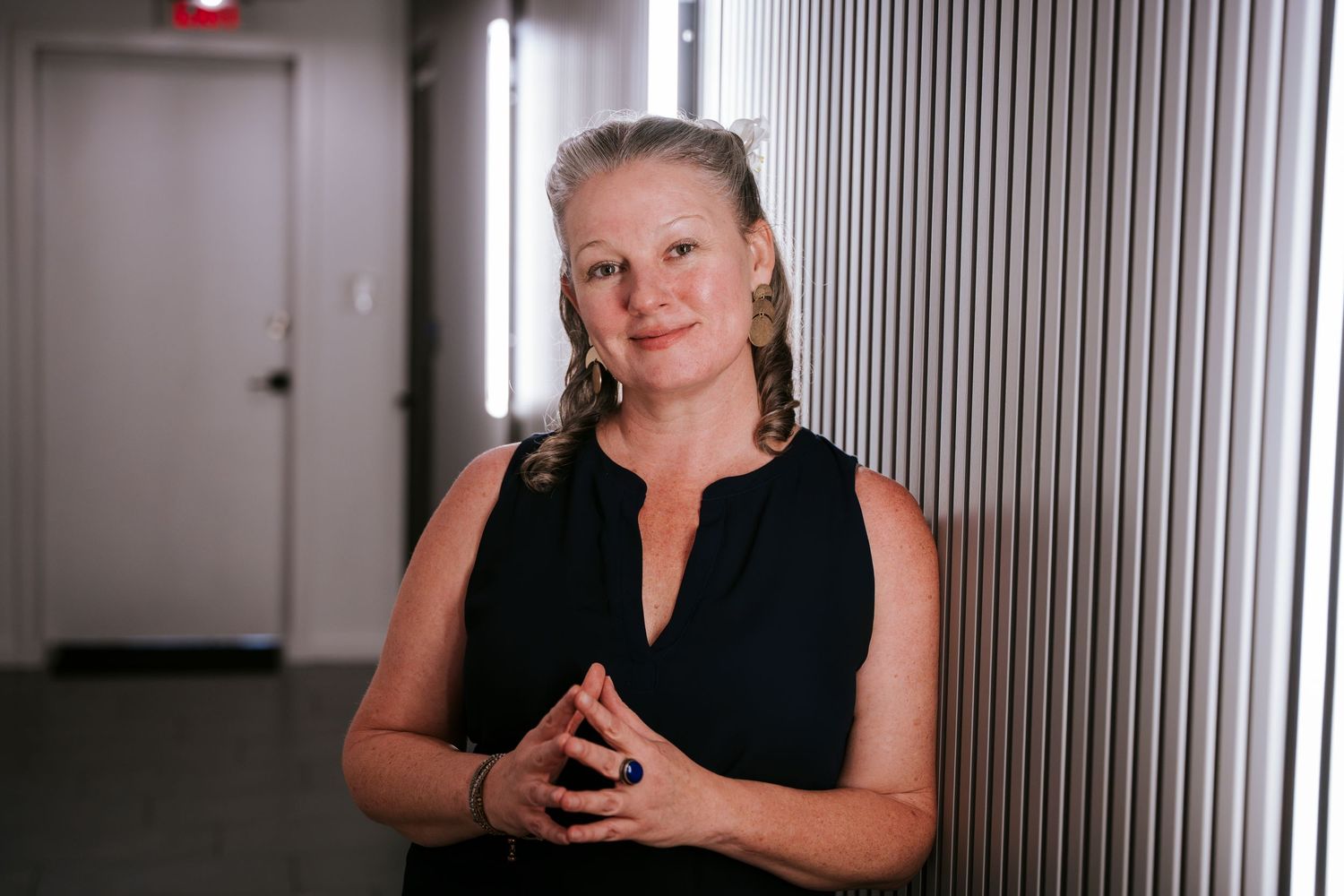 A person in a sleeveless black top stands in a corridor with metallic walls clasping their hands together.