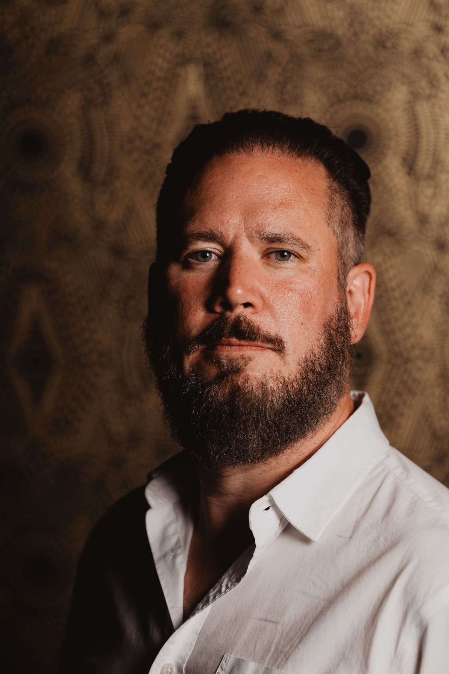 Professional studio portrait of a bearded man in a white button-down shirt against a textured brown background.