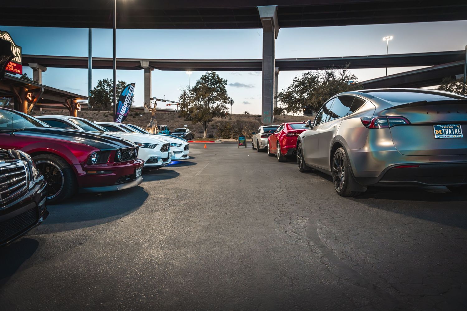 Row of luxury sports cars parked under an overpass at a car meet.