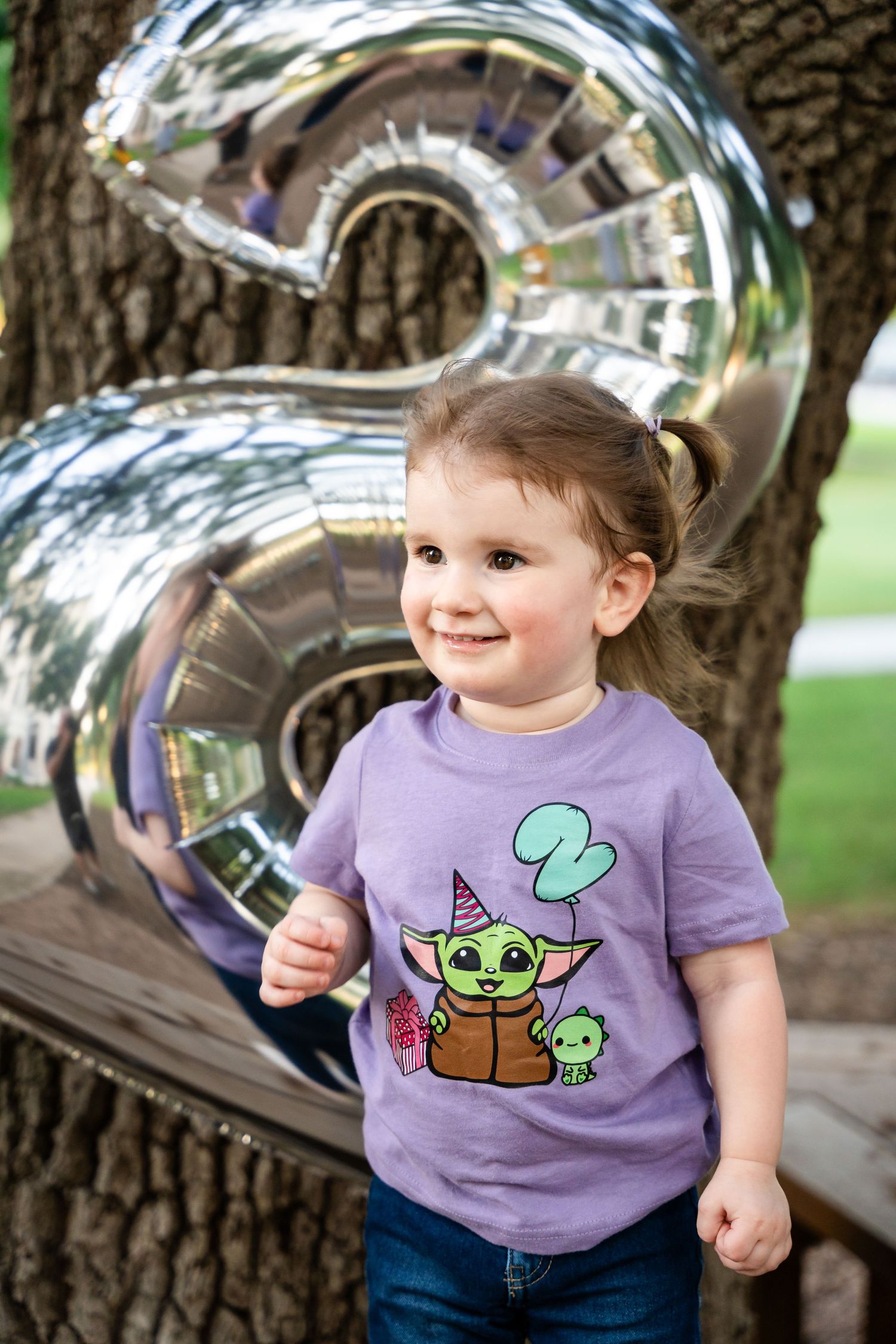 A purple t-shirt with Baby Yoda design is worn against a large silver number balloon backdrop in an outdoor setting.