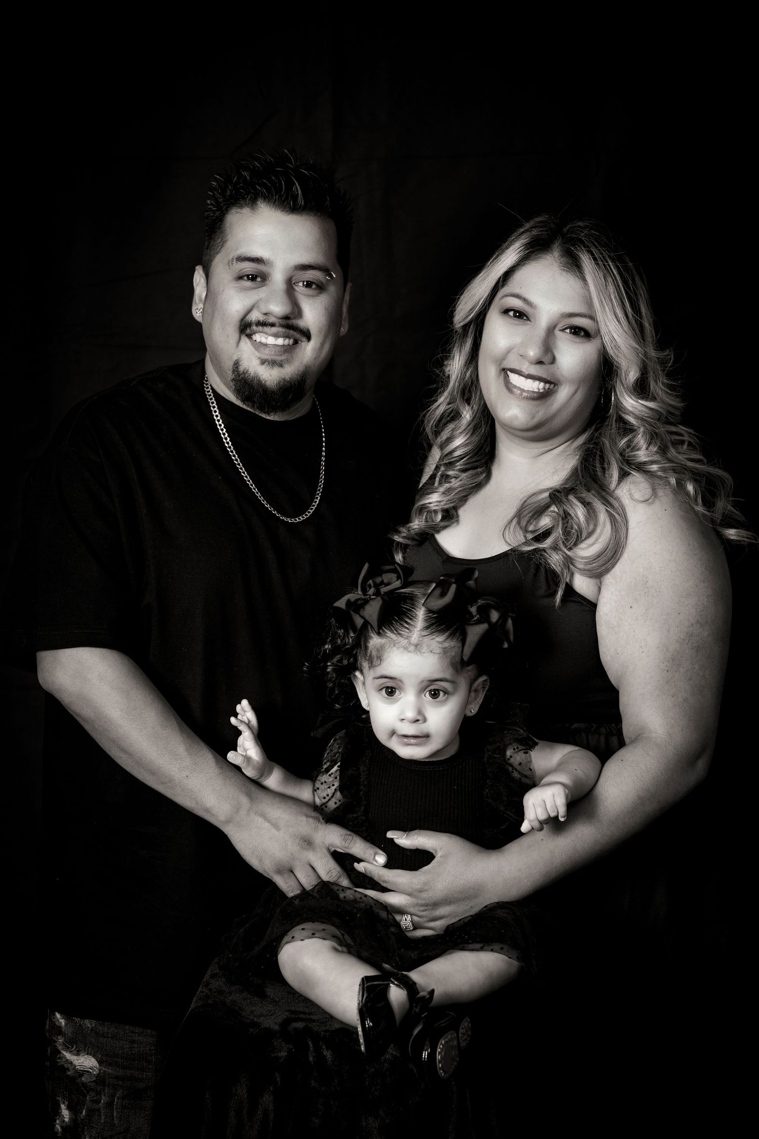 Black and white studio portrait of a family in matching dark outfits against dark background.