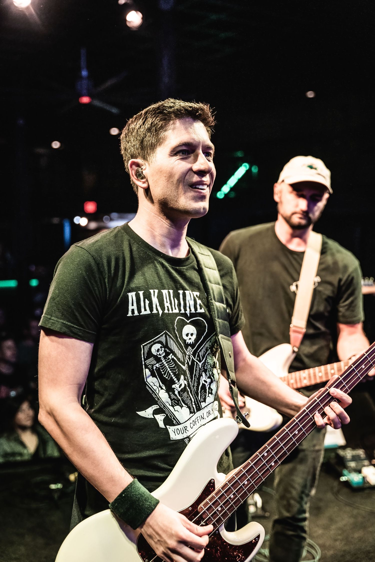 Musician plays bass guitar while performing on dark stage wearing Alkaline band t-shirt.