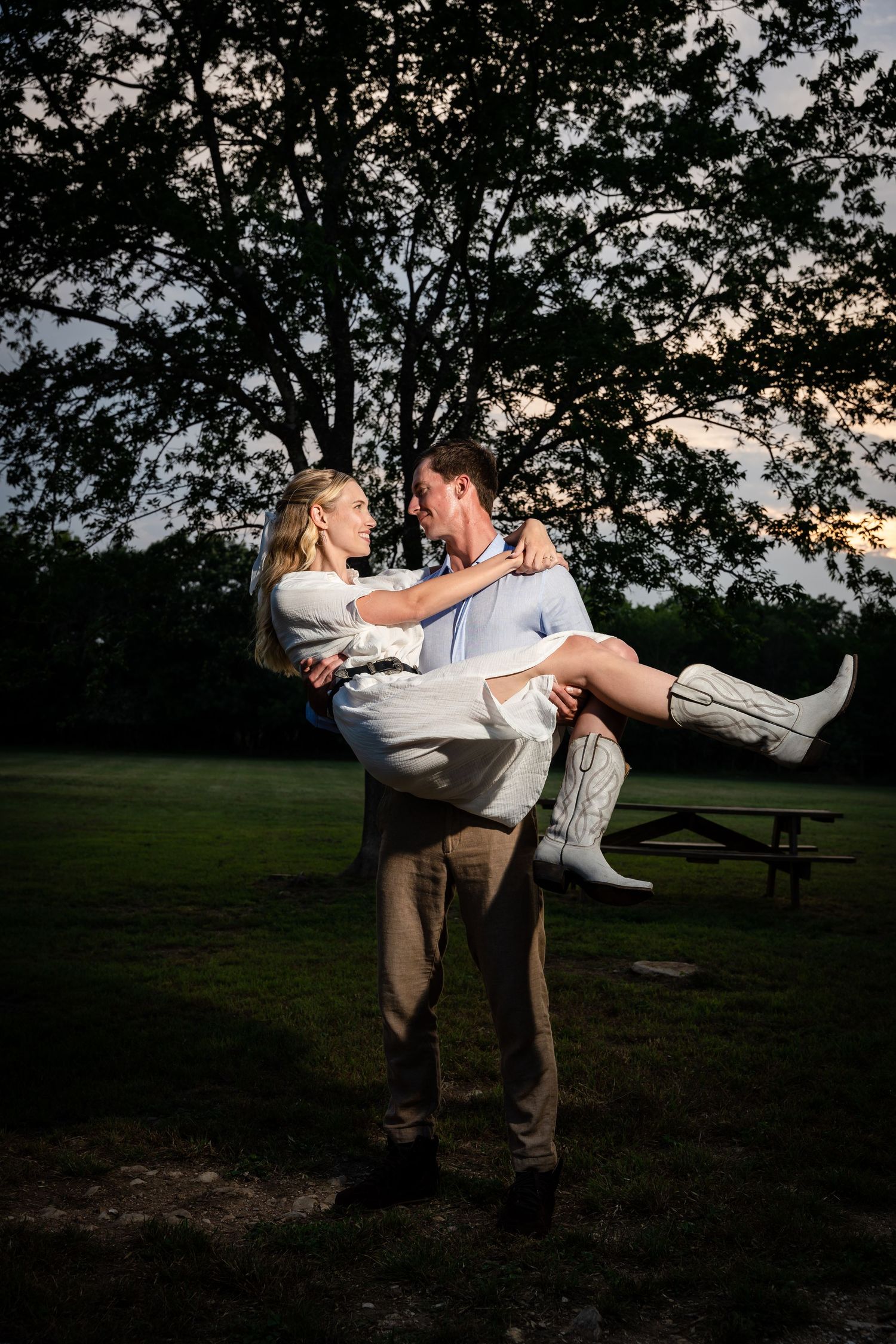 Couple shares a playful moment as one is lifted in white boots against a sunset sky.