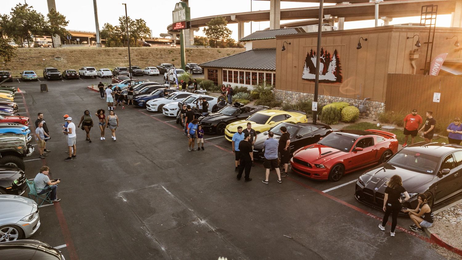Sunset view of car enthusiasts gathering around vehicles in a parking lot.