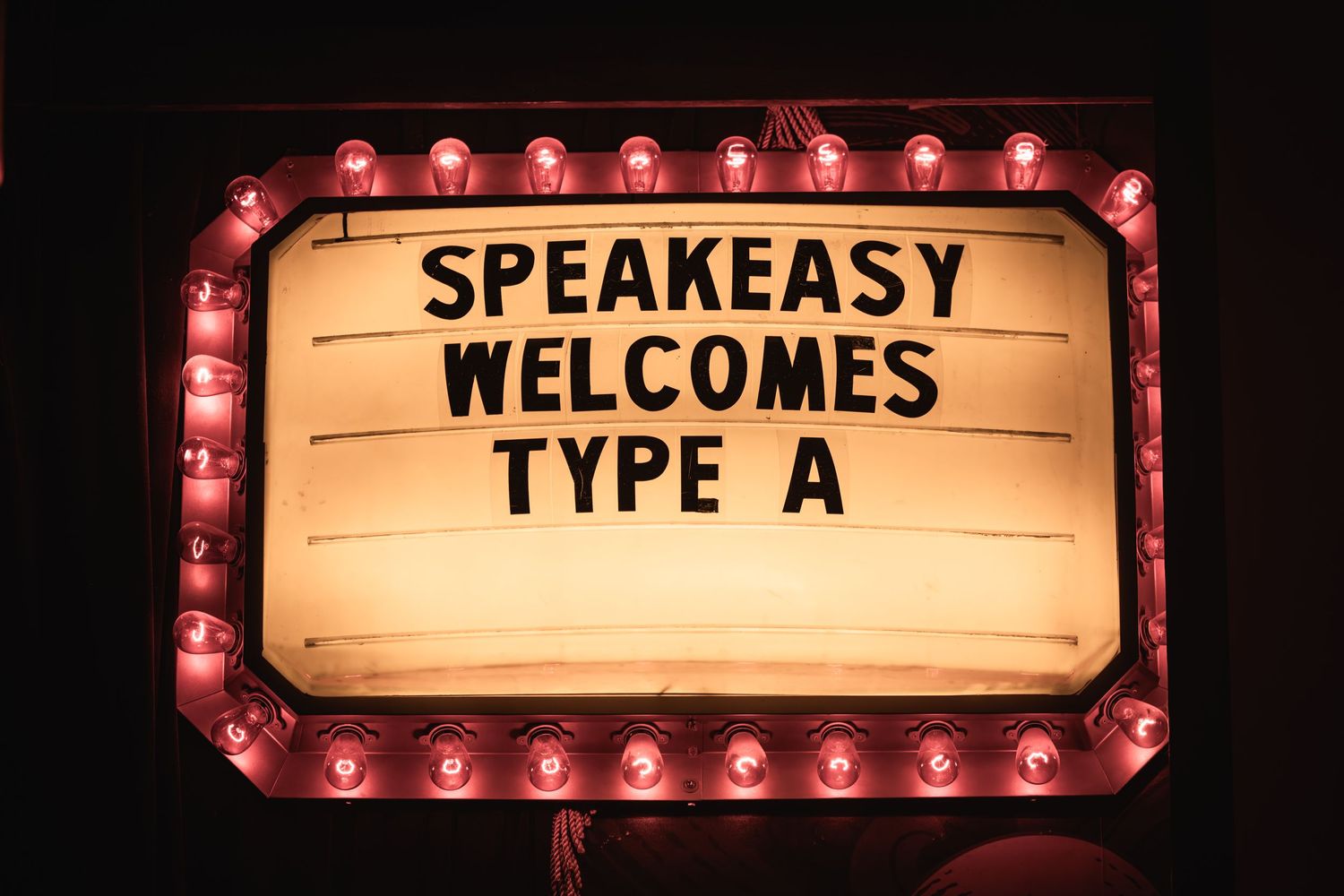 Illuminated vintage marquee sign reading 'SPEAKEASY WELCOMES TYPE A' with red light bulbs.