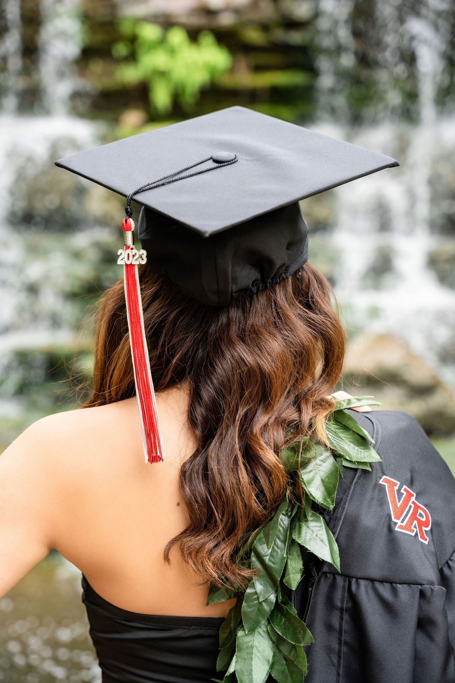Graduate wearing black cap with tassel and green leaf lei shown from behind near waterfall.