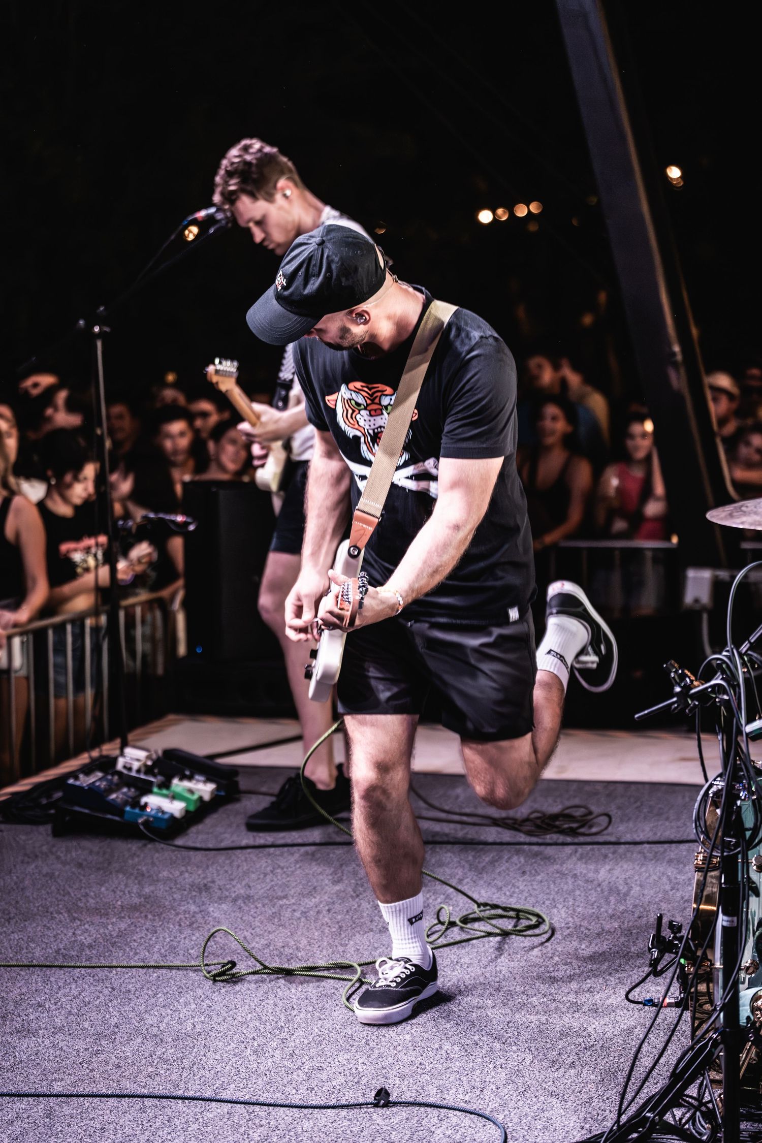 Musician playing guitar on stage with concert lighting and crowd barrier visible.