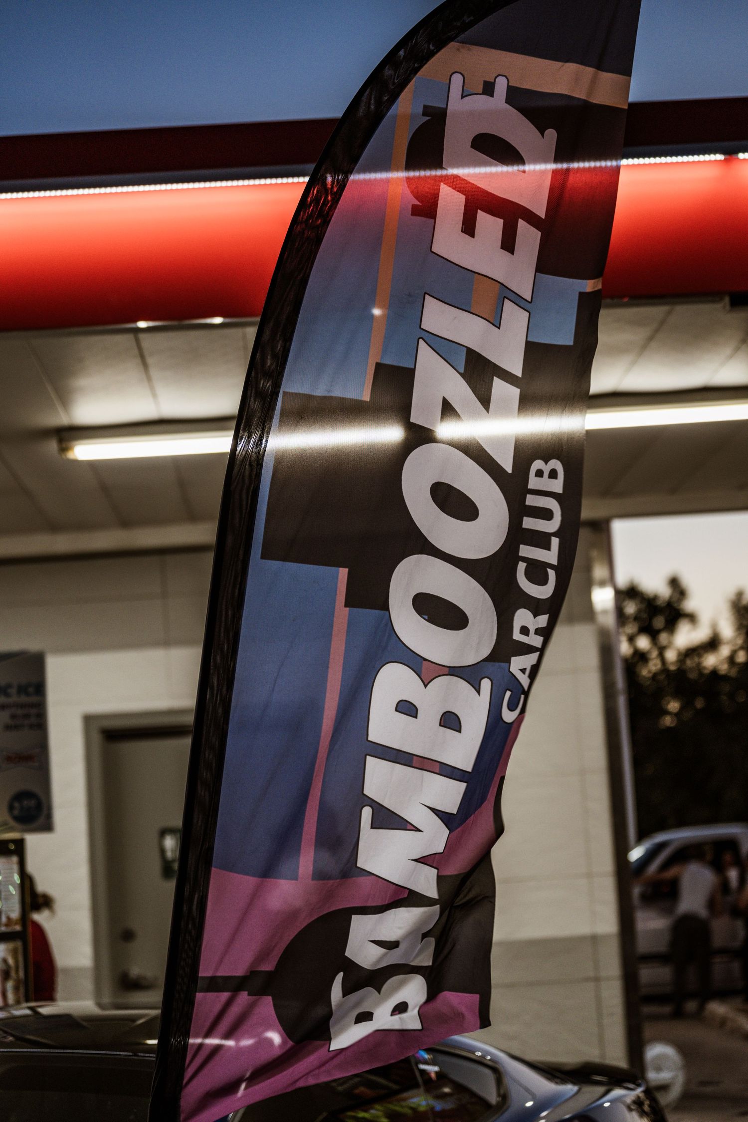 Bamboozer Car Club feather flag banner displayed against illuminated building exterior.
