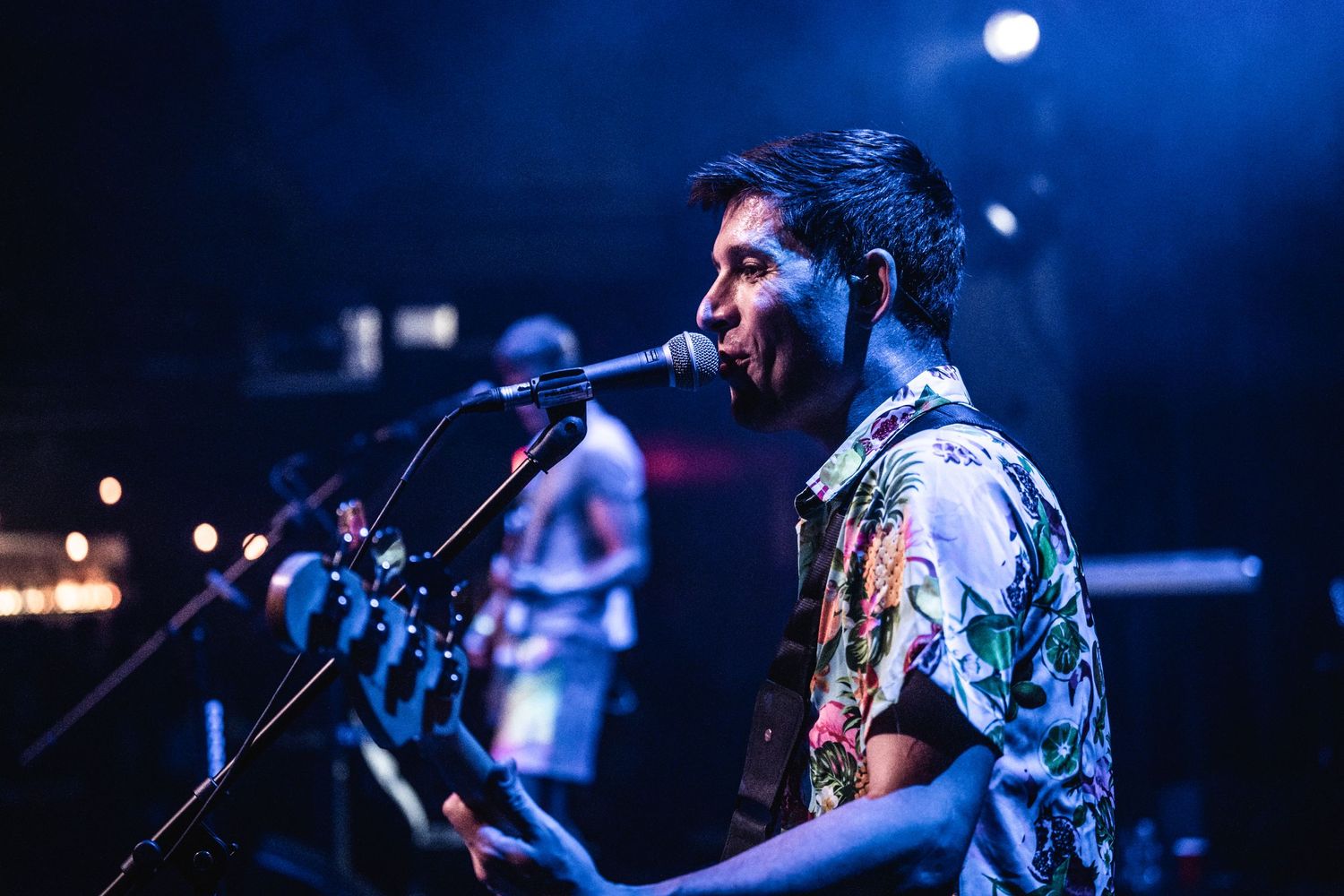 Musician performs at microphone in Hawaiian shirt against blue stage lighting at concert.