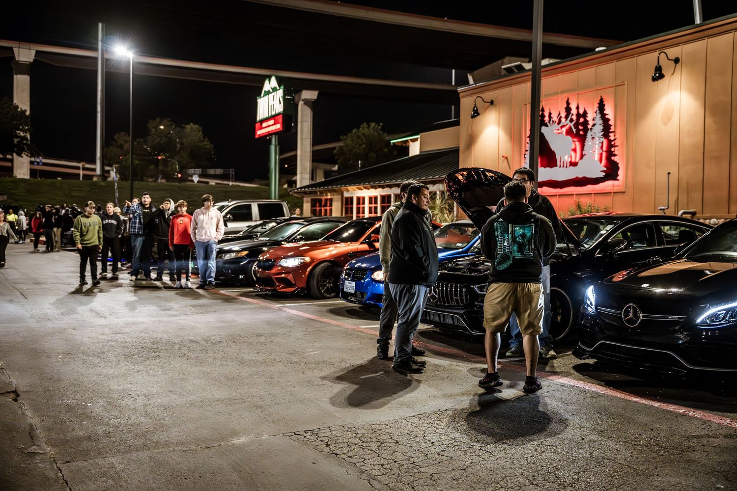 Crowd gathered at nighttime car meet outside retail shopping center.