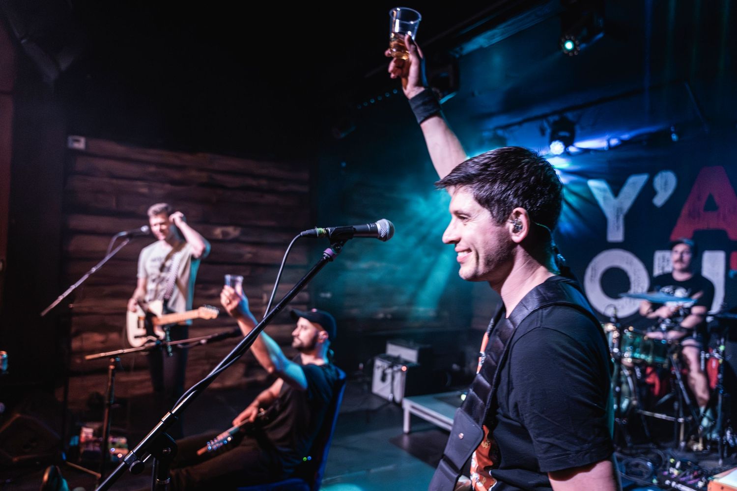 Rock band performs energetically on stage with colorful blue lighting and multiple microphones.