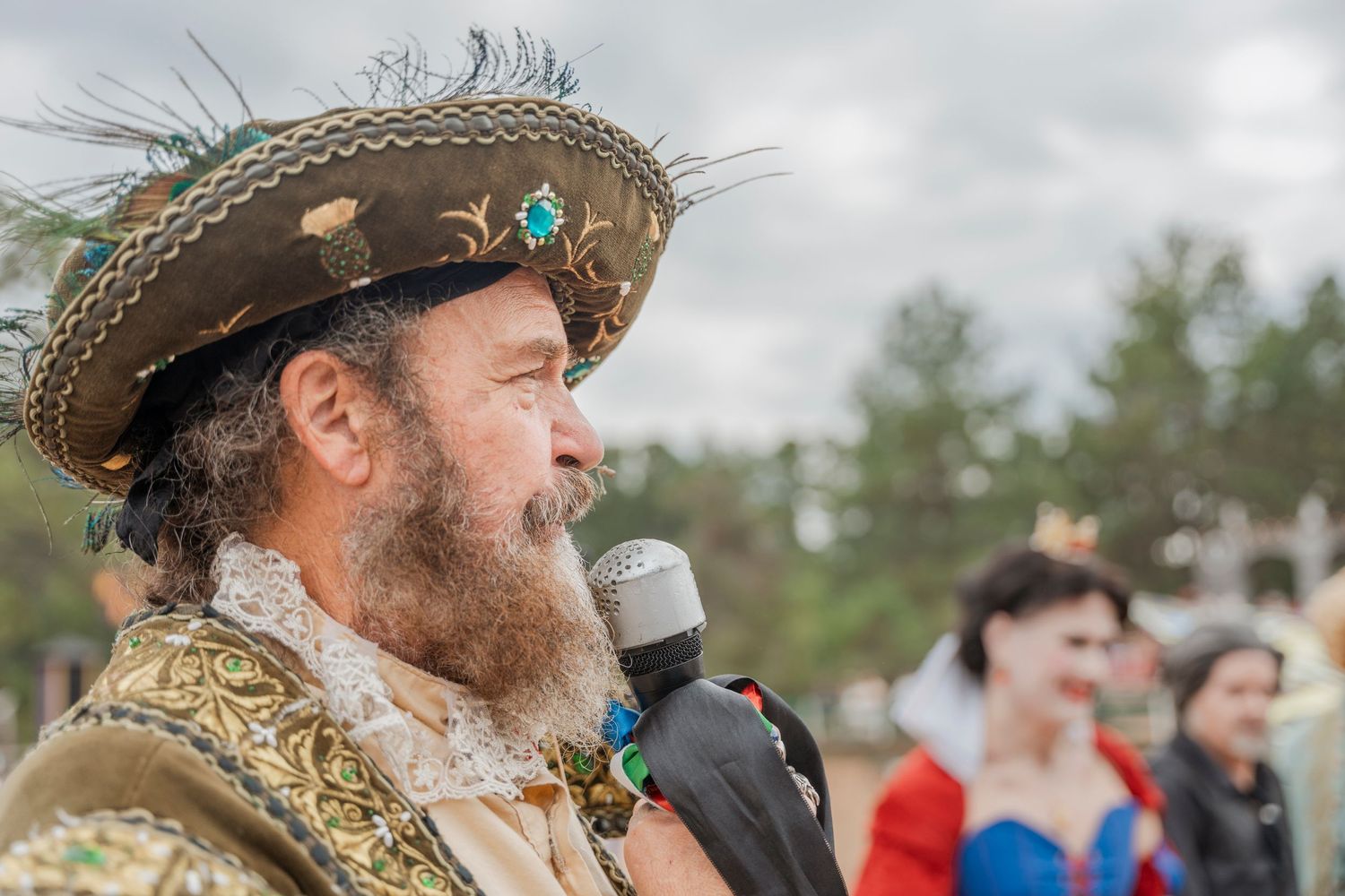 Medieval costumed performer wearing decorated straw hat speaks into microphone at renaissance faire.