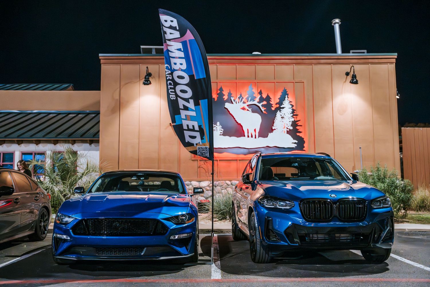Two blue performance cars parked under promotional flags outside a building at night.