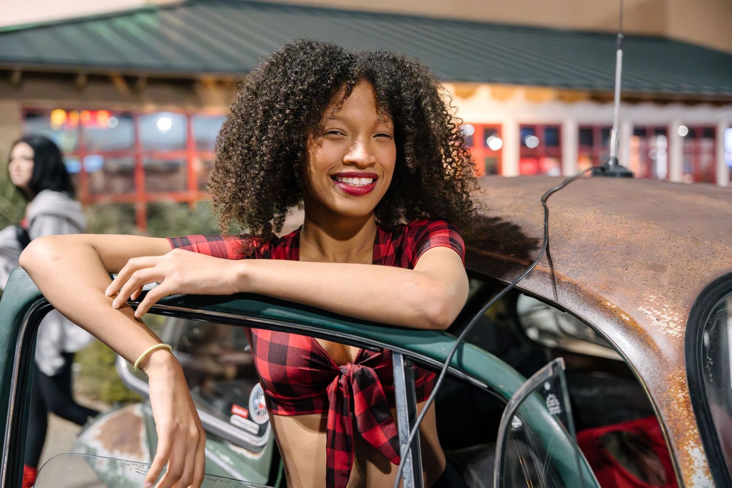 Person in red plaid top leaning on vintage car door with bright smile.