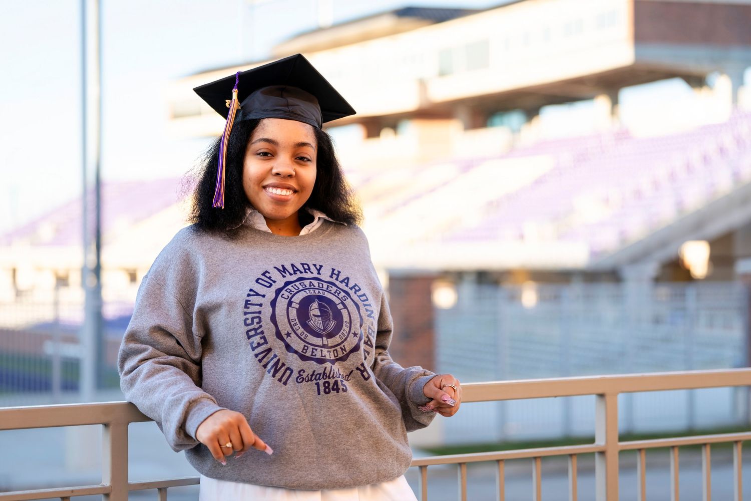 A graduate in university sweatshirt and cap poses near stadium bleachers during golden hour.