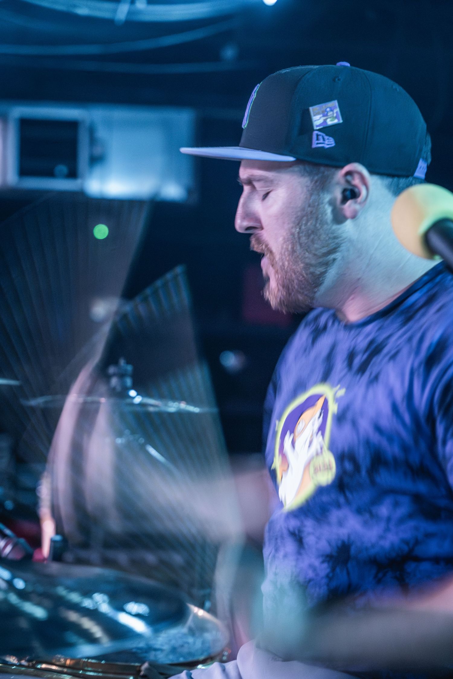 Drummer performing on stage in a baseball cap and blue shirt with dramatic concert lighting.