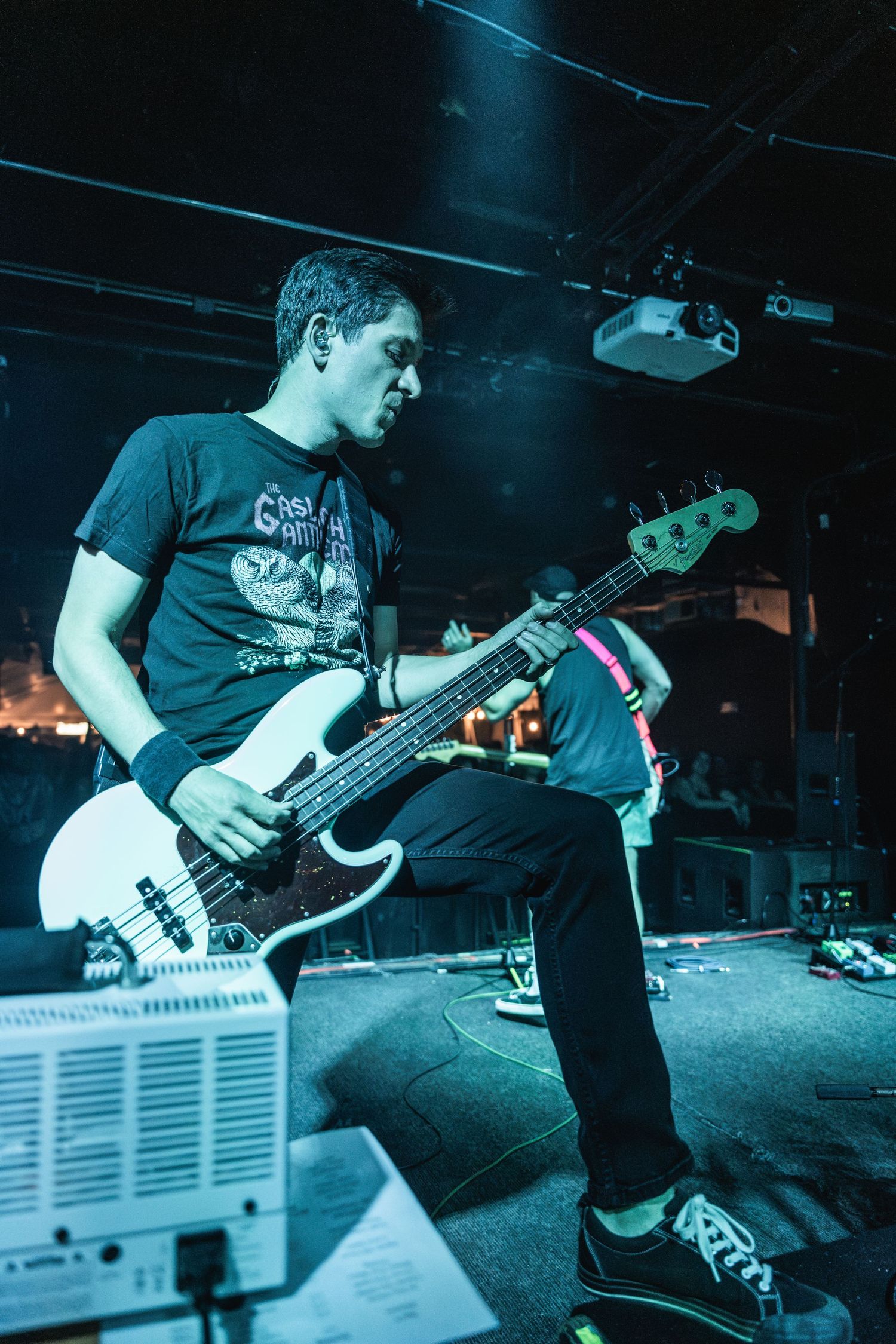 A bassist in a black t-shirt plays on stage under dramatic blue lighting.