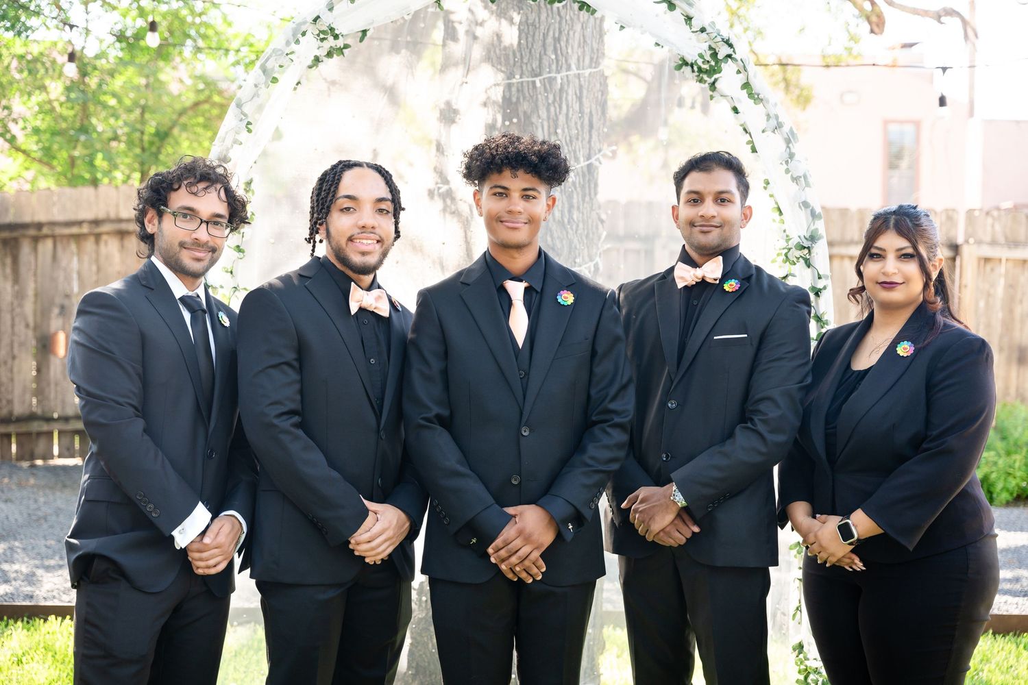 Wedding party members in black formal attire stand in a row under a garden arch.