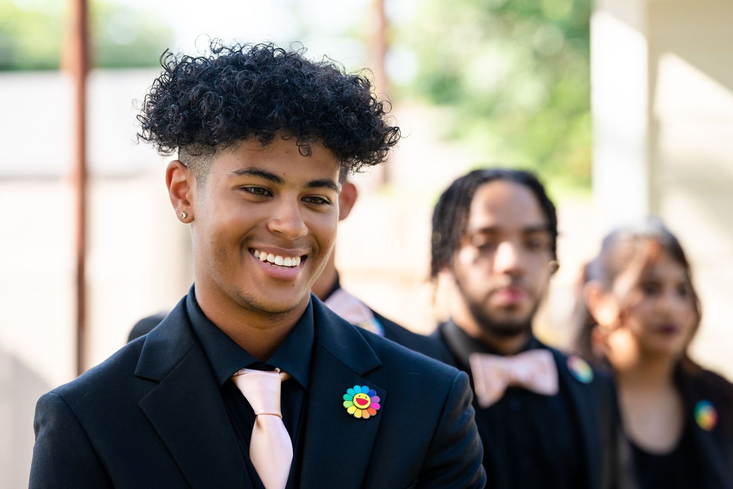 A person in formal attire wearing a pink tie and rainbow pin smiles during a celebration.