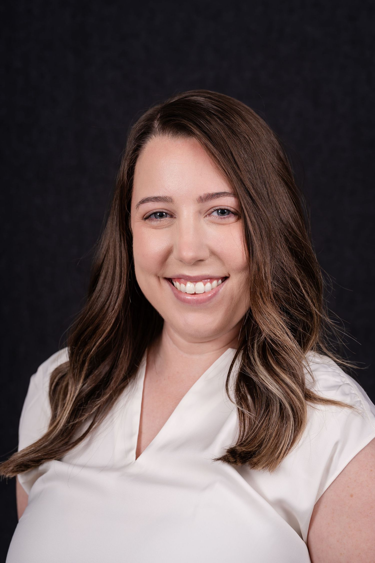 Professional headshot of a smiling person with long brown wavy hair wearing a white blouse against a dark background.