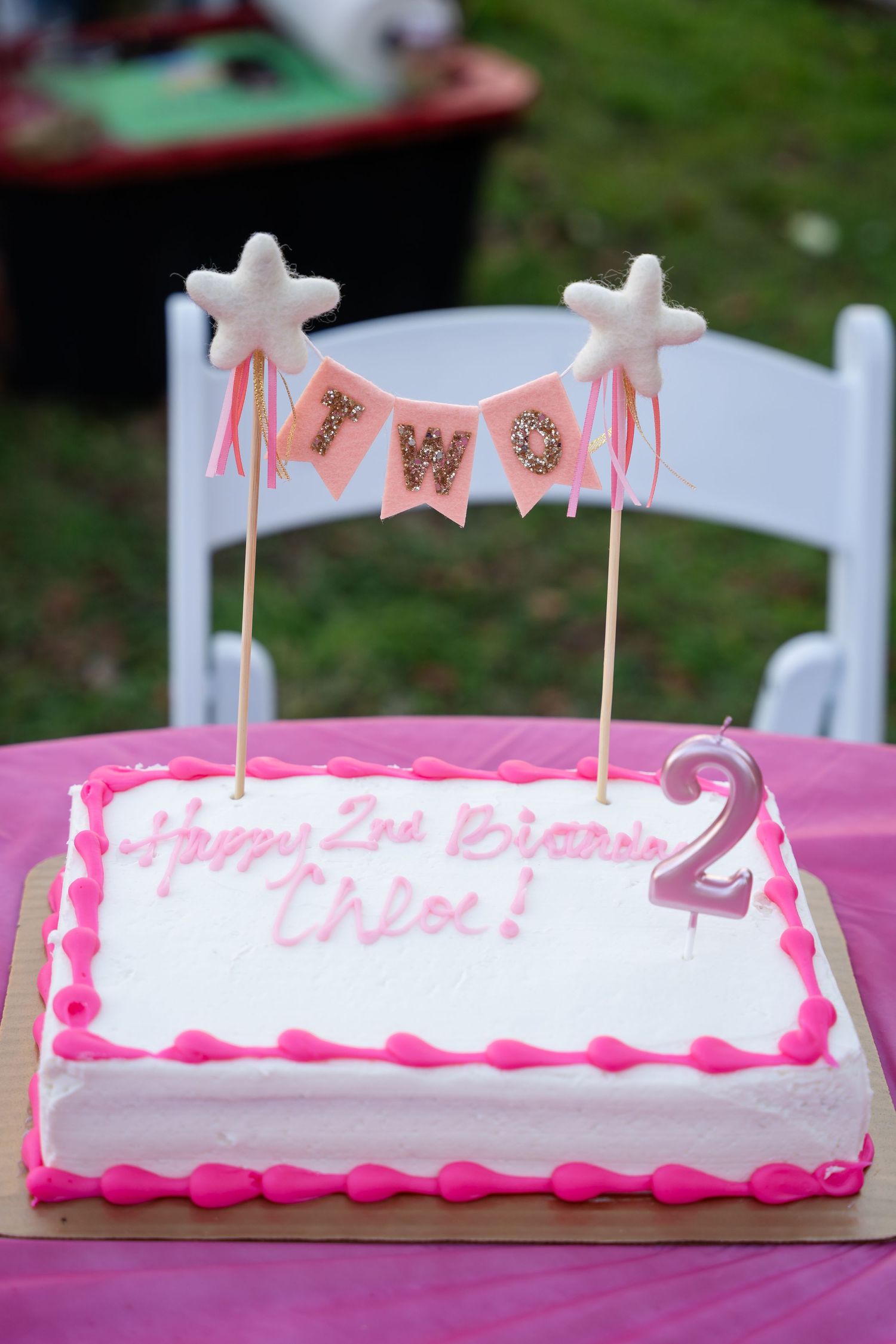 White birthday cake with pink trim and banner decoration featuring stars on sticks and a number 2 candle on top.