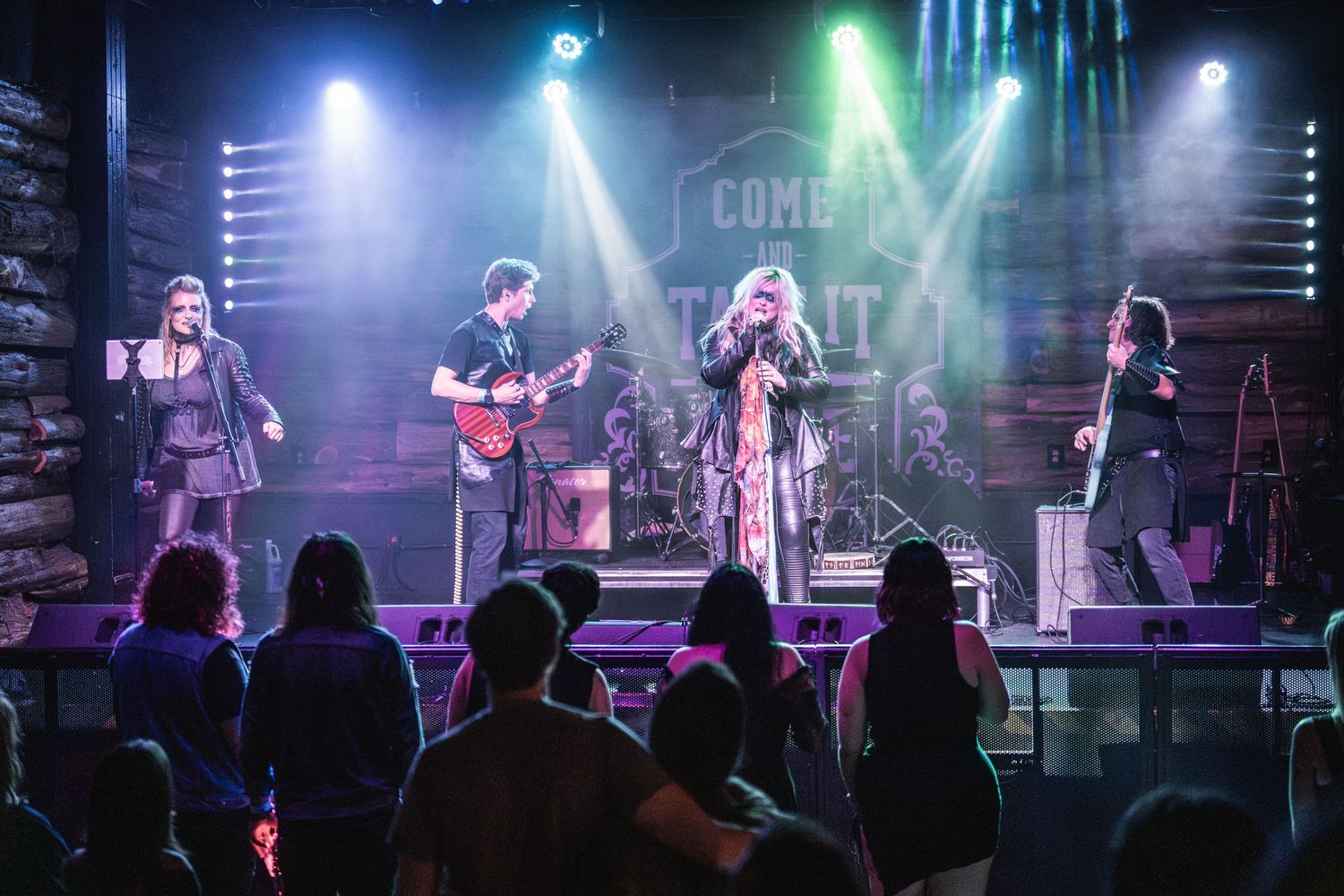 Full band performs on elevated stage with blue lighting and audience silhouettes visible.
