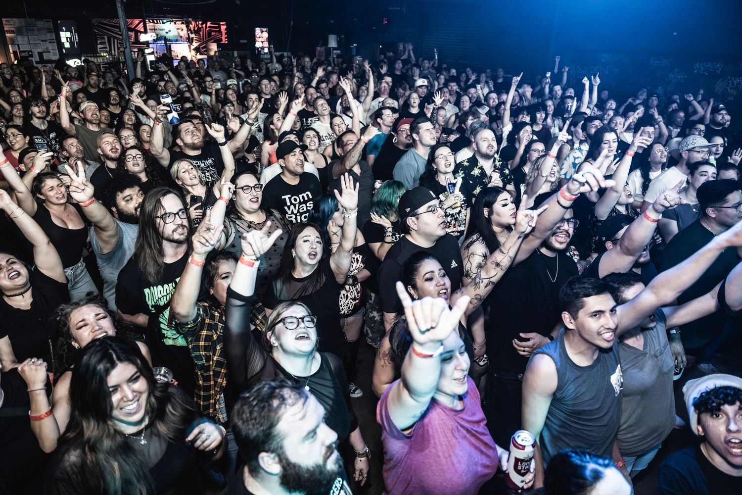 Large energetic crowd of concert goers with raised arms enjoying a music performance in a dark venue with blue lighting.