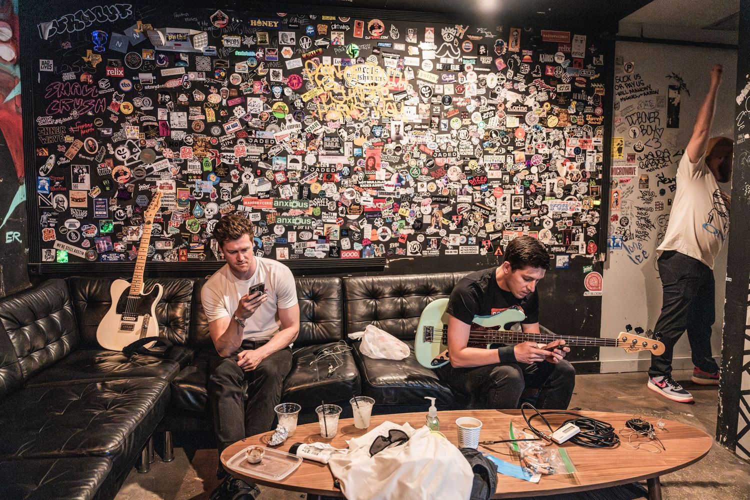 Musicians relax in a small basement room with guitars and equipment, surrounded by countless photos covering the walls.