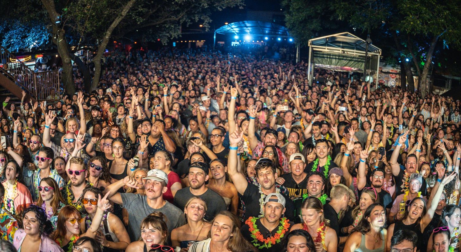 Aerial view of a massive outdoor concert crowd with raised arms under stage lights at night.