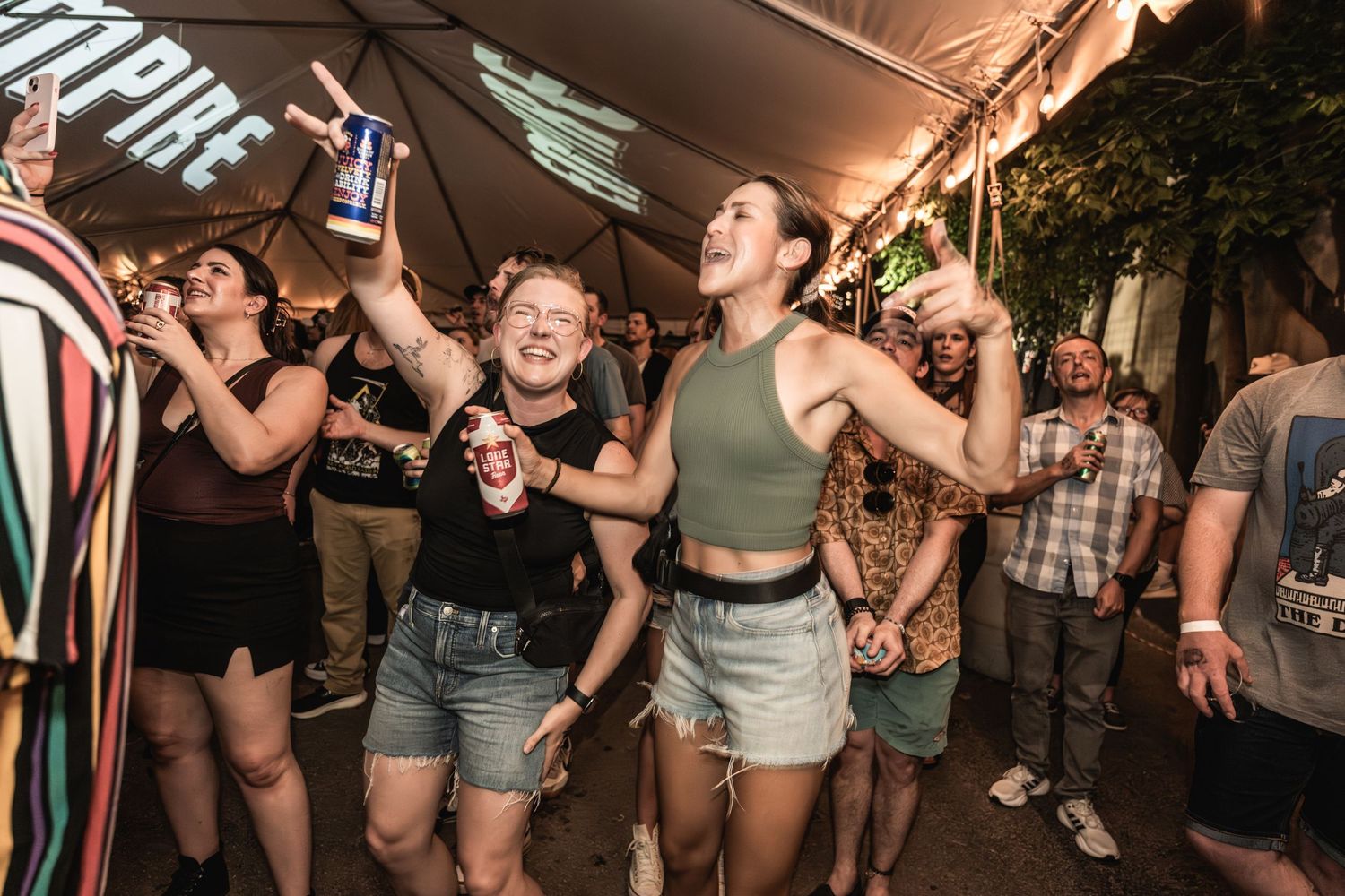 Party attendees celebrate and raise drinks during an outdoor event under string lights.