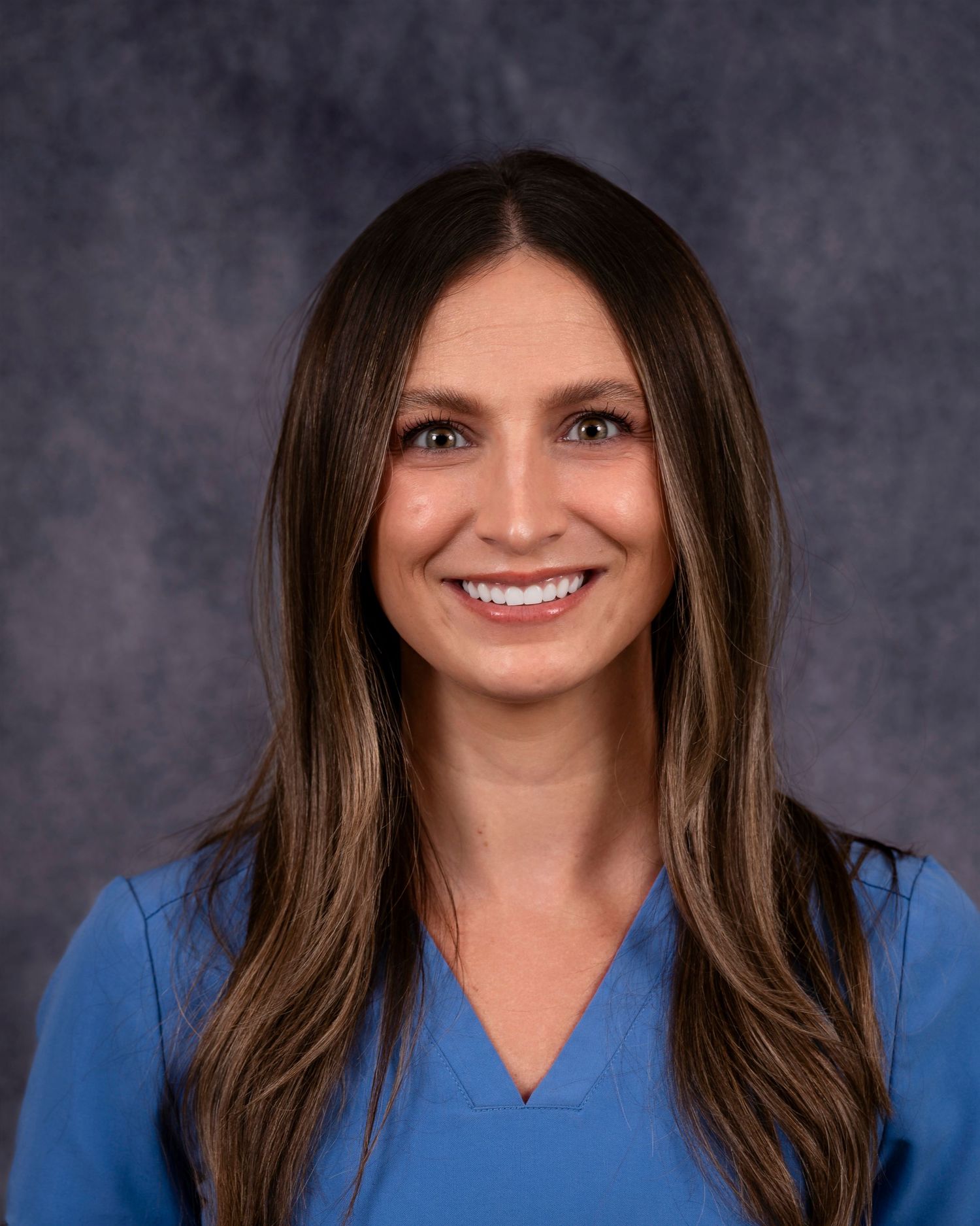 Professional portrait of someone with long brown hair wearing a blue v-neck top against a dark gray background.