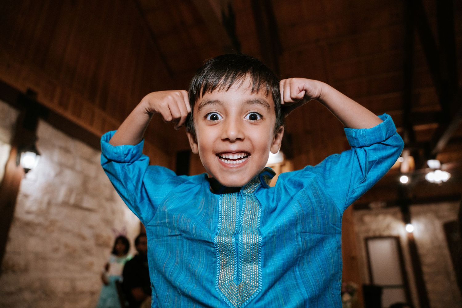 A joyful child in a bright blue traditional outfit makes a playful expression at a celebration.