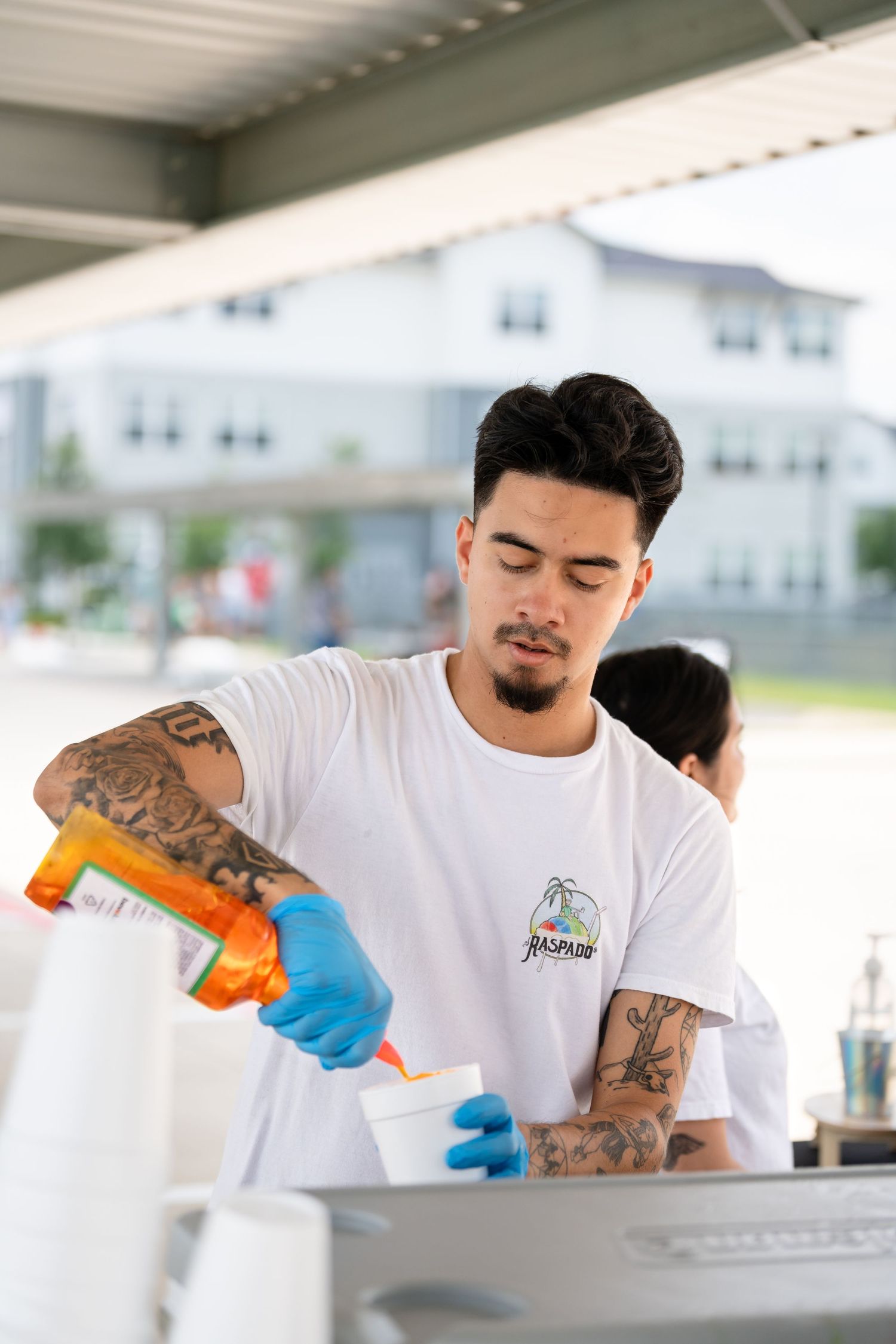 Tattooed person in white t-shirt pouring a beverage while wearing blue gloves.