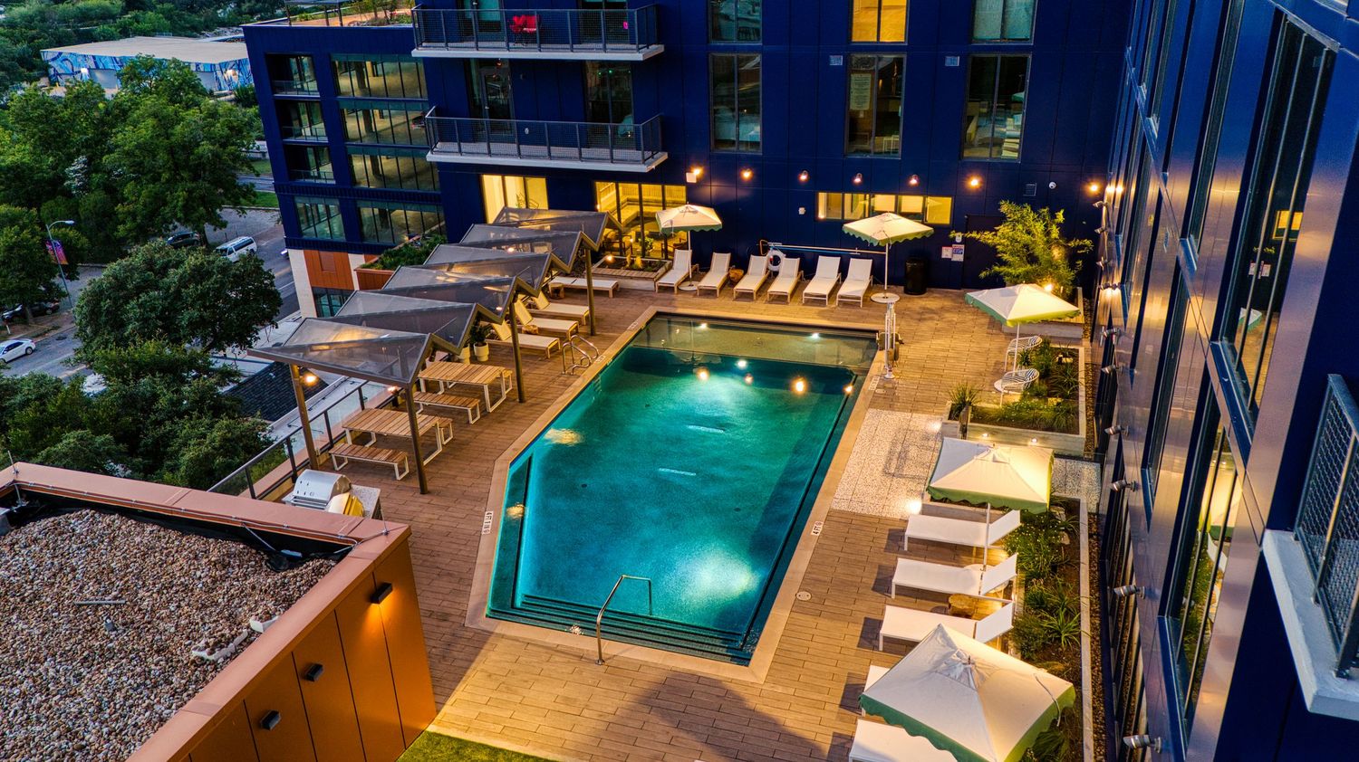 Evening view of illuminated rooftop pool deck with lounge chairs and dining areas at luxury apartment complex.