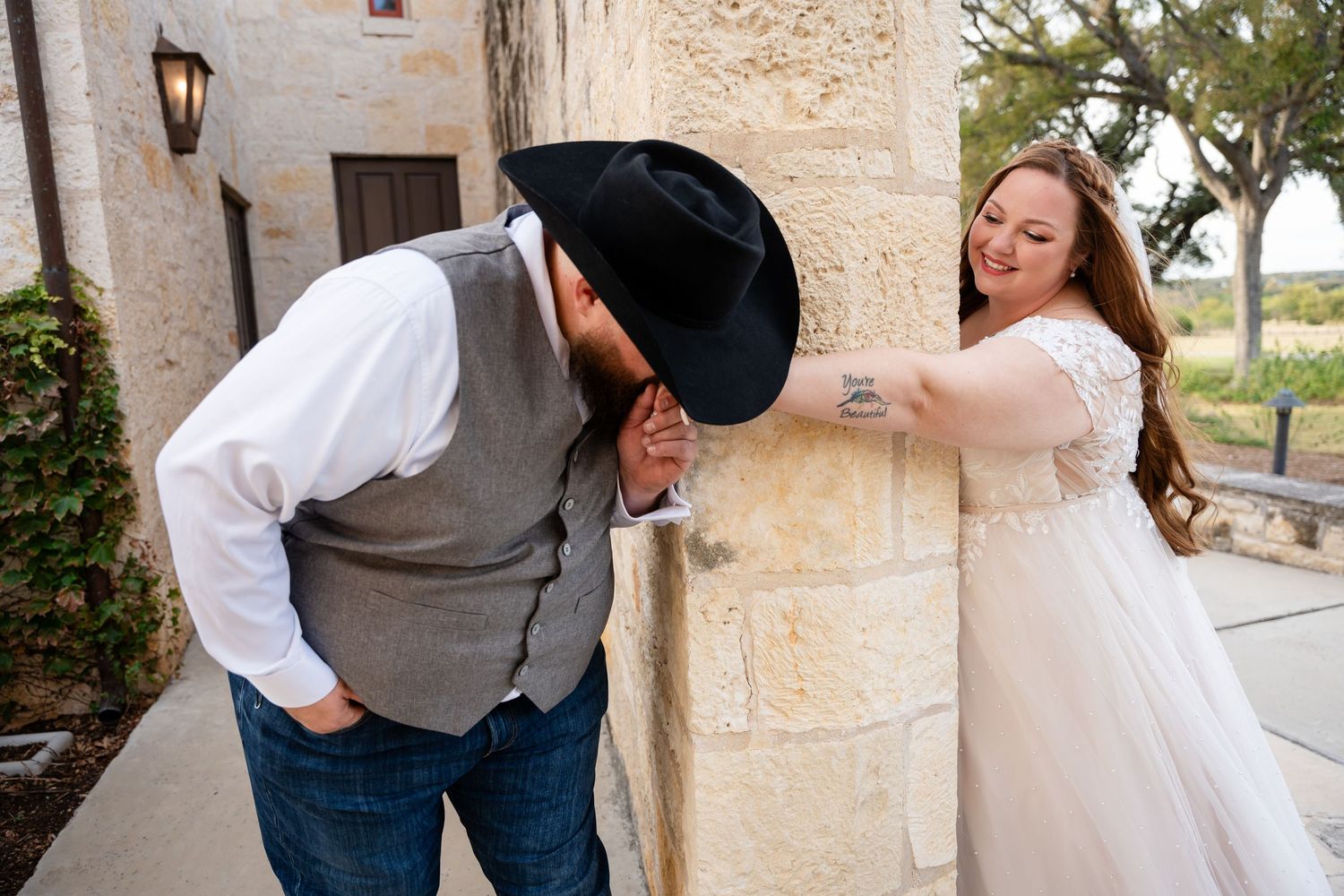 A playful couple in western wedding attire share a fun moment against a stone pillar.