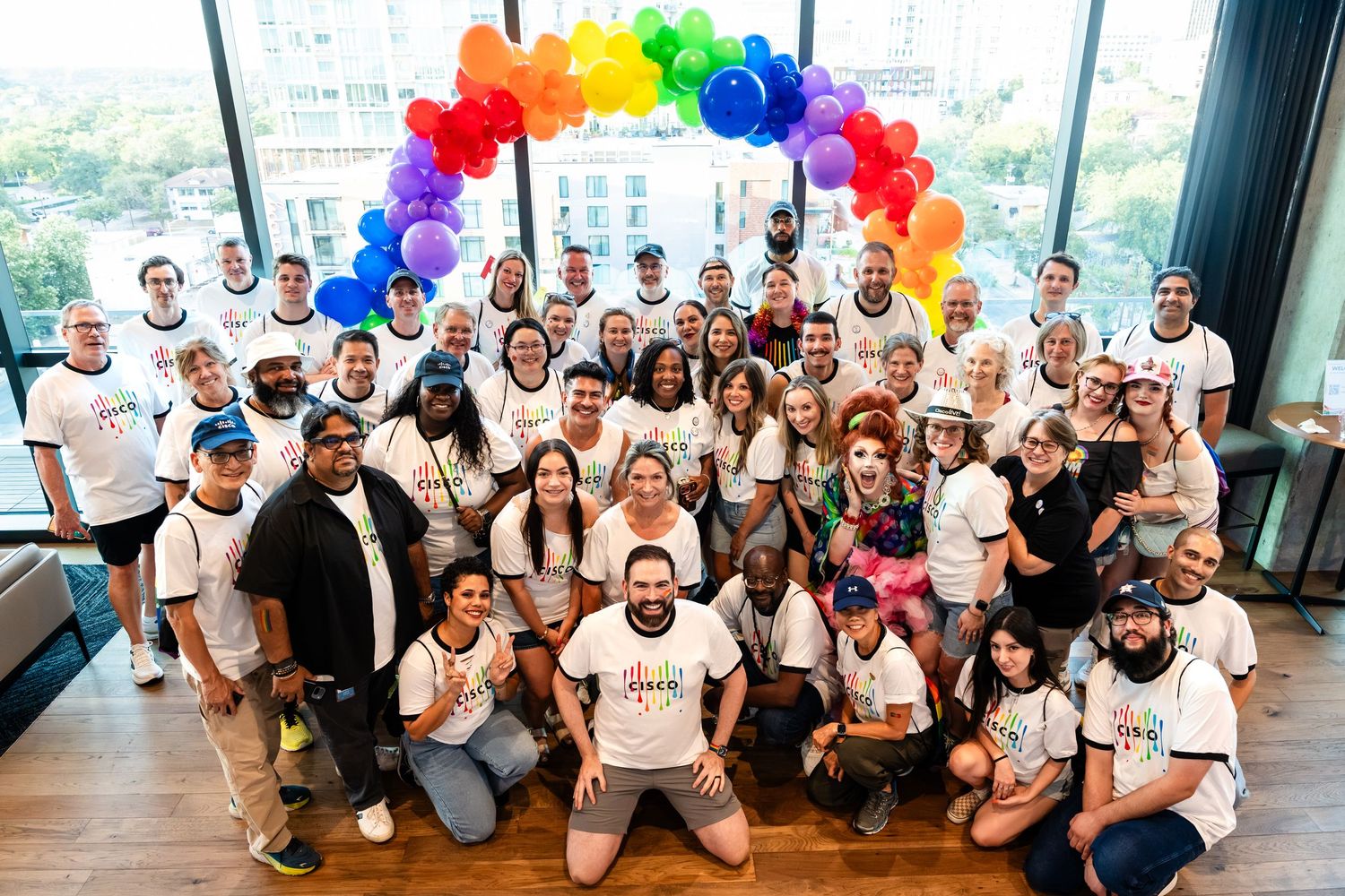 Large group poses under rainbow balloon arch in white Pride t-shirts at indoor celebration event.
