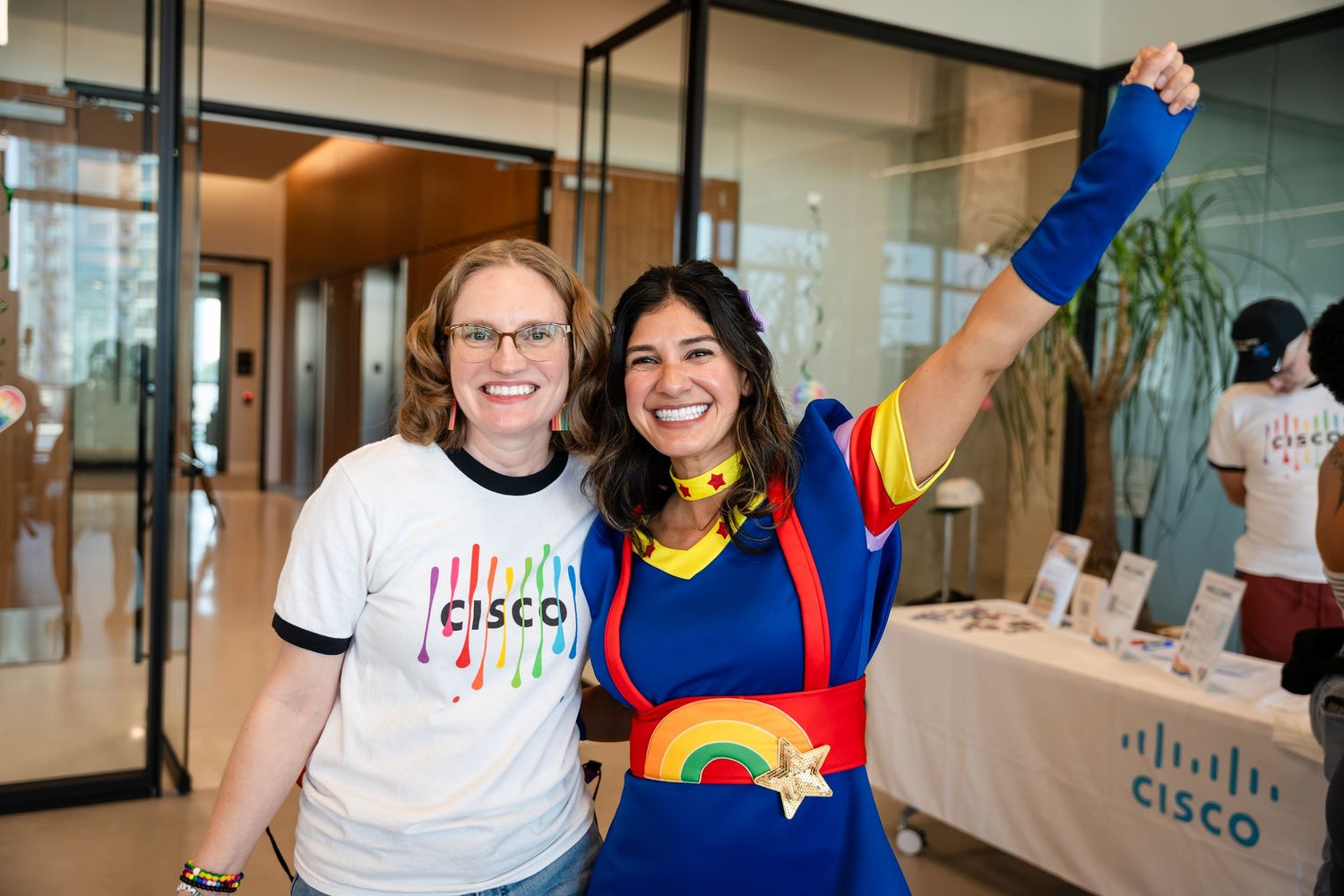 Two women smiling at a Cisco event, one in a rainbow Cisco shirt, the other in a colorful superhero costume.