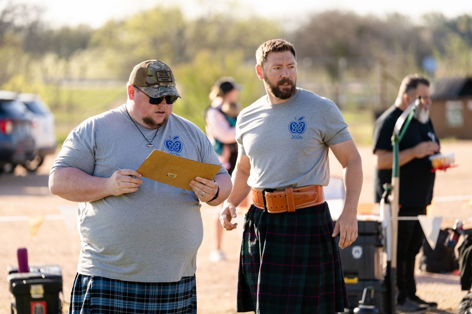 Two men in kilts and gray shirts at a Highland Games event, one reviewing documents outdoors.