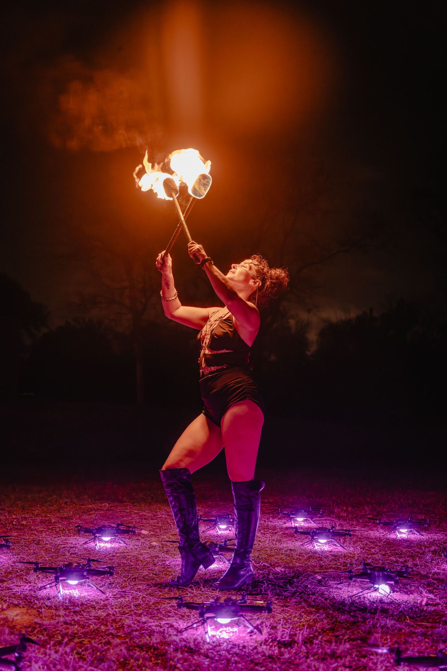 Fire performer in black outfit and boots twirls flaming baton at night, surrounded by purple ground lights.
