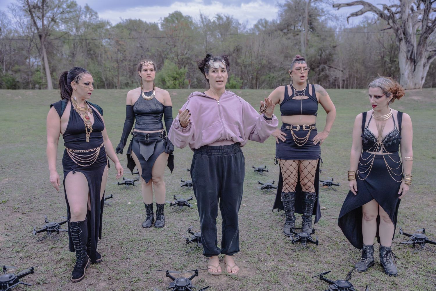 Five women in dramatic outfits stand in a field surrounded by drones during an outdoor performance.