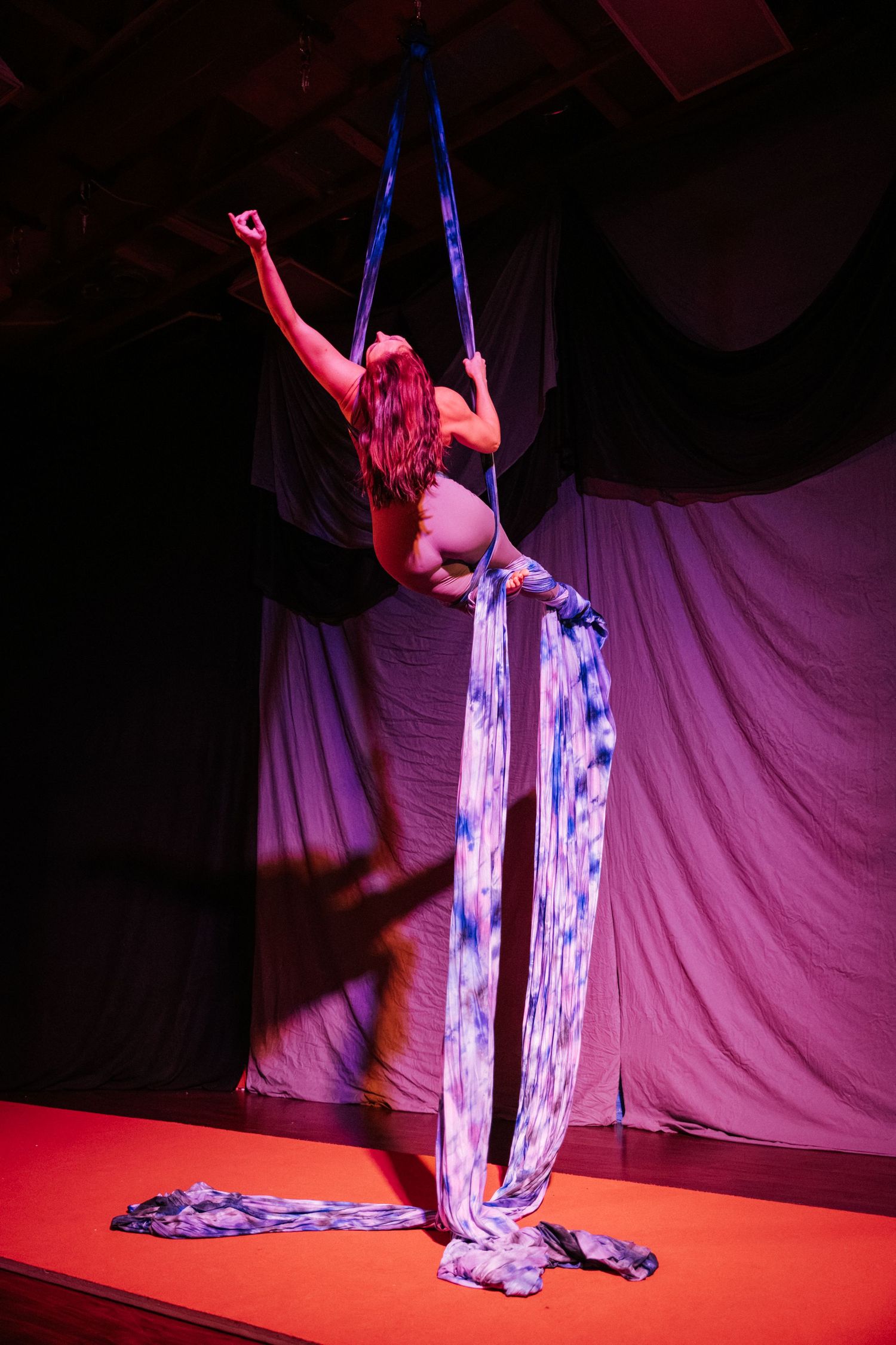 Aerial silk performer on stage with blue tie-dye fabric under dramatic red and purple lighting.
