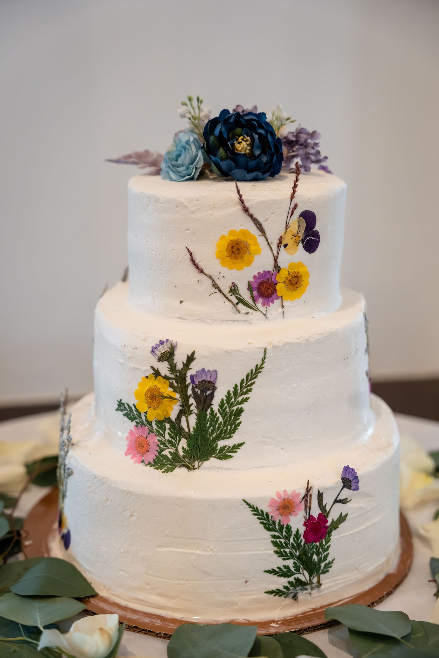 Three-tier white wedding cake decorated with pressed wildflowers and topped with blue and purple blooms on wooden stand.