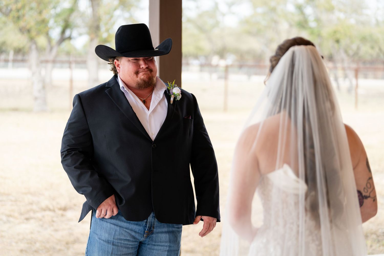 Groom in black cowboy hat and blazer with jeans shares first look moment with bride in white wedding dress and veil.
