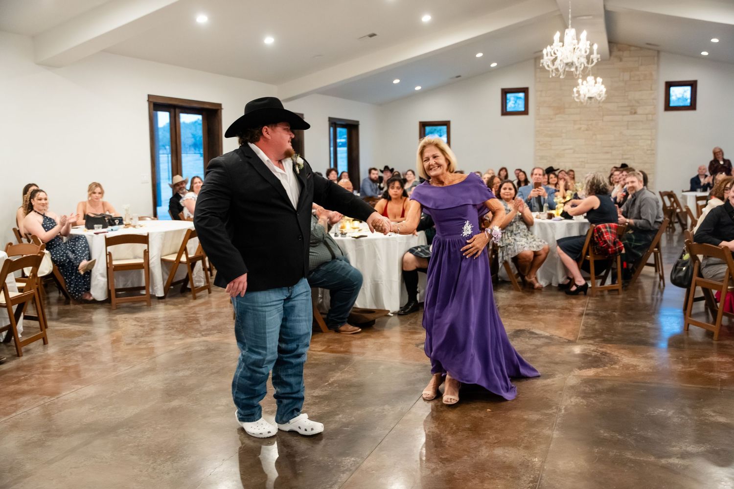 Couple dancing in elegant wedding venue with guests seated at tables in rustic stone ballroom with chandelier lighting.