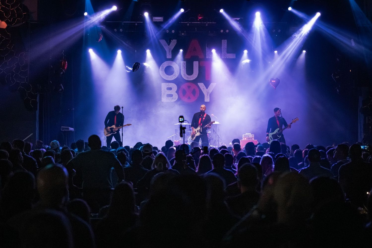 Band performing on stage with dramatic blue and white spotlights before a large crowd at a You Boy concert.